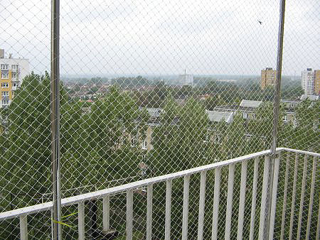 Balkon zabezpieczony siatką ochronną przed ptakami, widok na miasto zza siatki, biała balustrada.